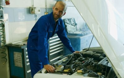 A smiling mechanic in a blue overall inspecting a car engine in a garage.
