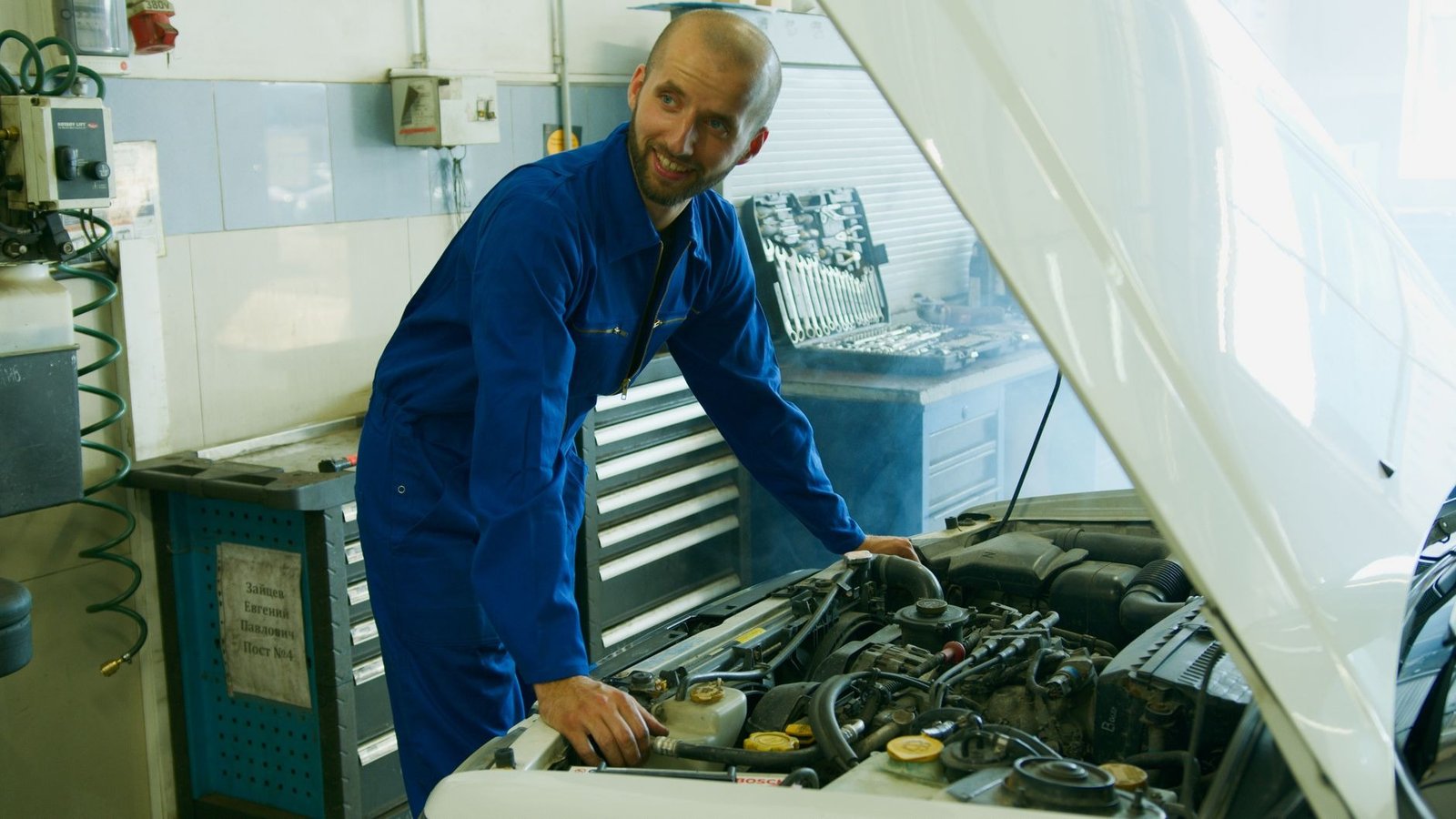 A smiling mechanic in a blue overall inspecting a car engine in a garage.