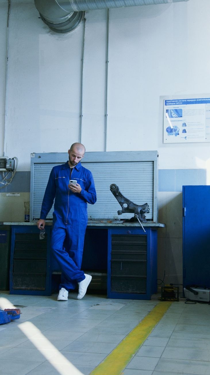 Mechanic wearing blue jumpsuit stands indoors using phone in repair shop.