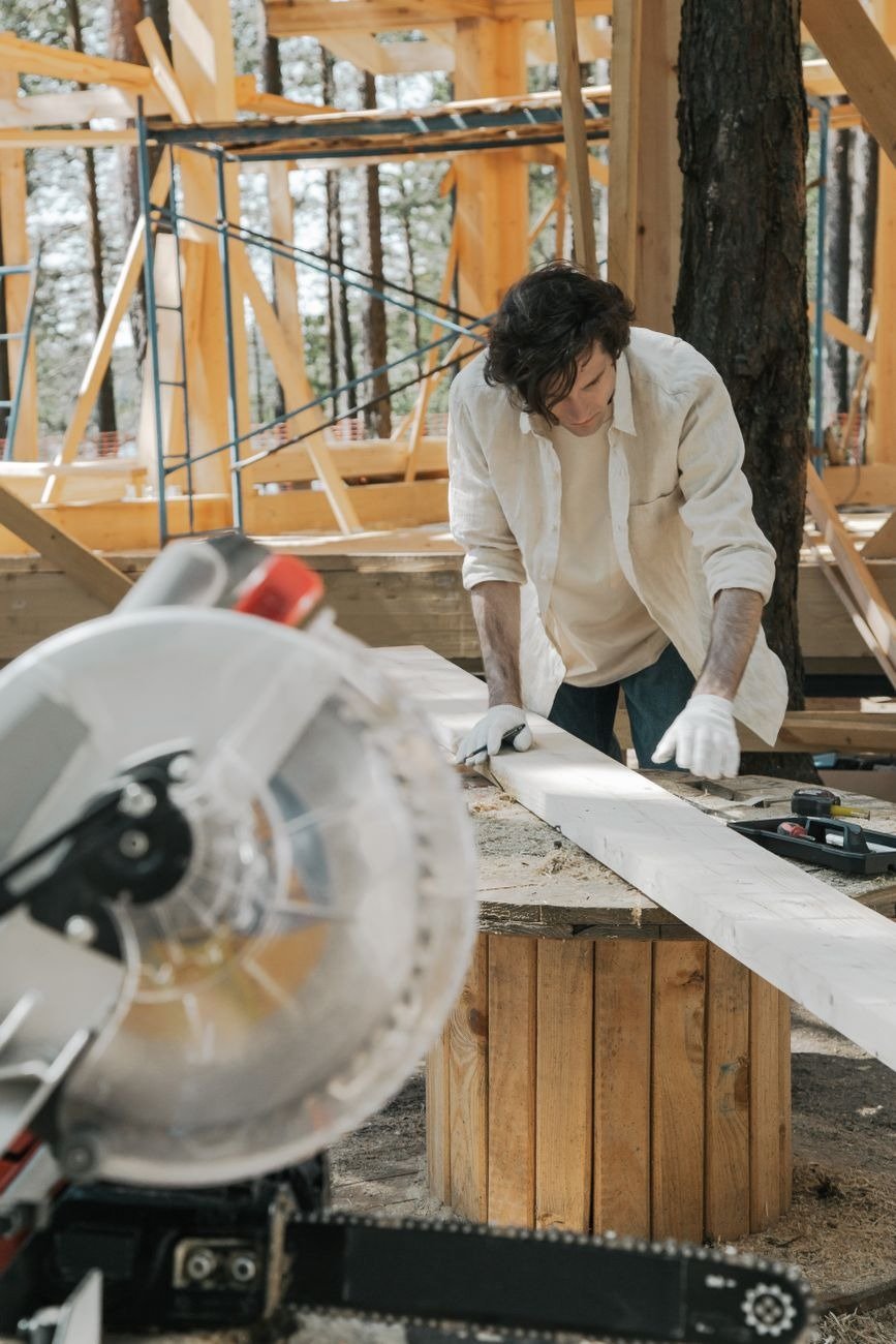 A carpenter uses a circular saw on a wood plank in an outdoor construction setting.