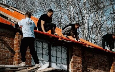 Workers repairing a brick building roof surrounded by birch trees on a clear day.