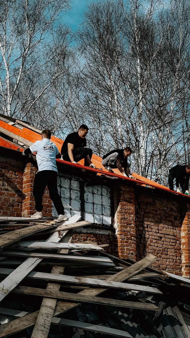 Workers repairing a brick building roof surrounded by birch trees on a clear day.