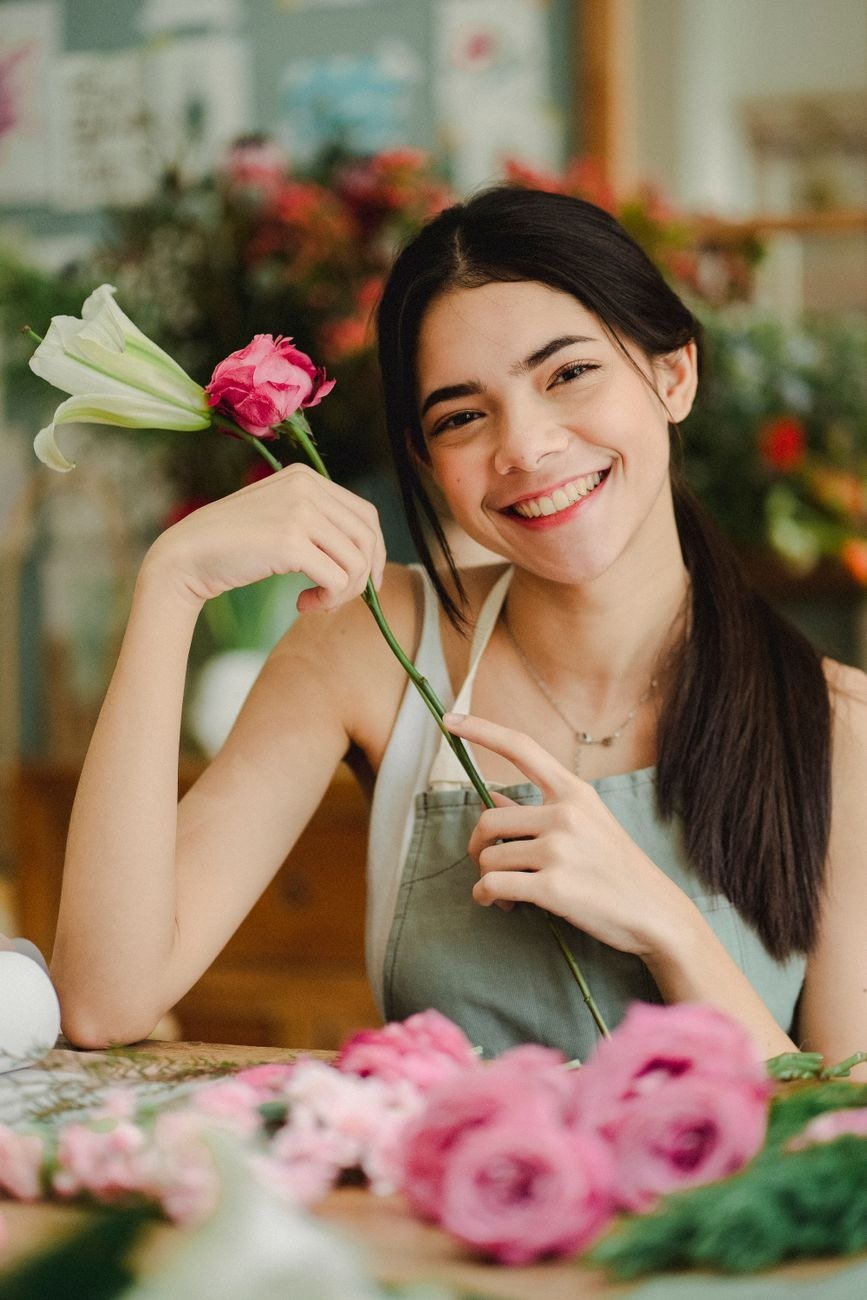 Young cheerful female worker with toothy smile making floral composition with roses and lilies in flower store