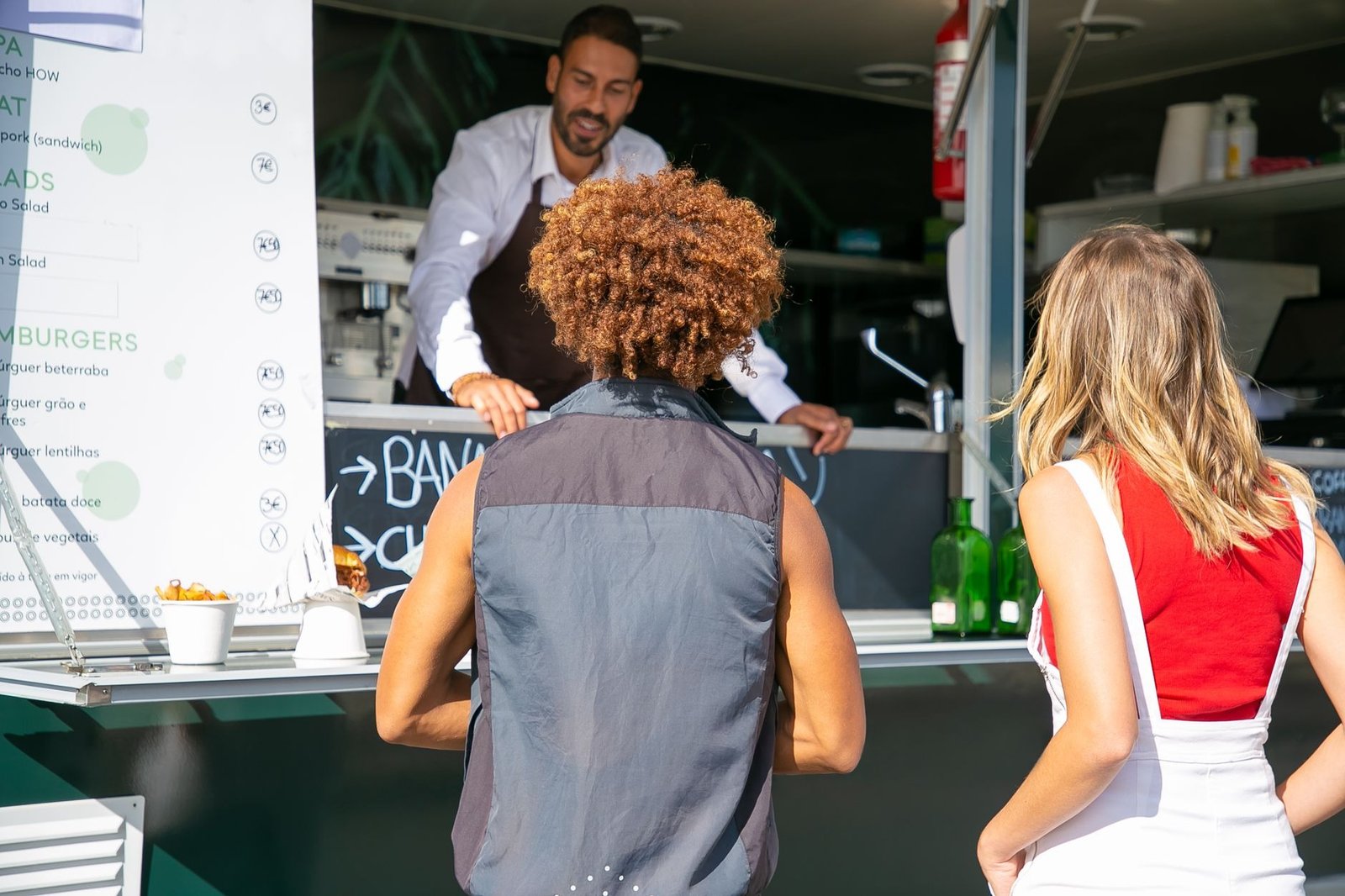 Hospitable ethnic man in apron standing at counter and helping diverse customers with choice while working in food truck