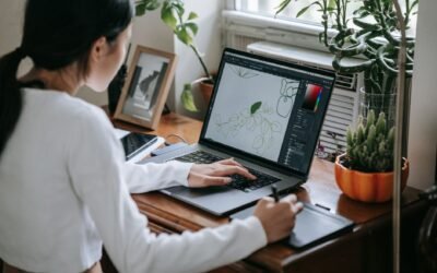 Woman using a stylus to draw on a laptop at a desk, surrounded by plants indoors.