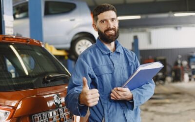 Auto mechanic in blue coveralls with clipboard giving thumbs up in garage.