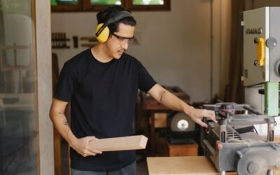 Young serious ethnic male woodworker in protective headphones and glasses with timber touching electric equipment outdoors