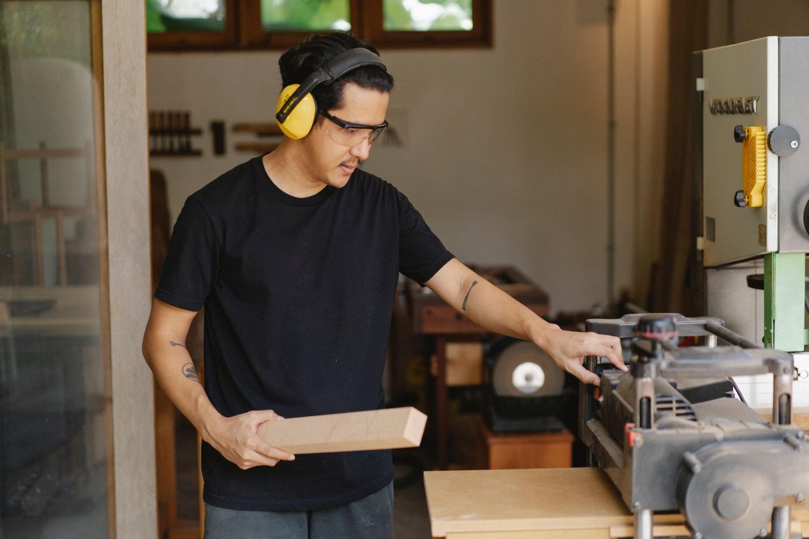 Young serious ethnic male woodworker in protective headphones and glasses with timber touching electric equipment outdoors