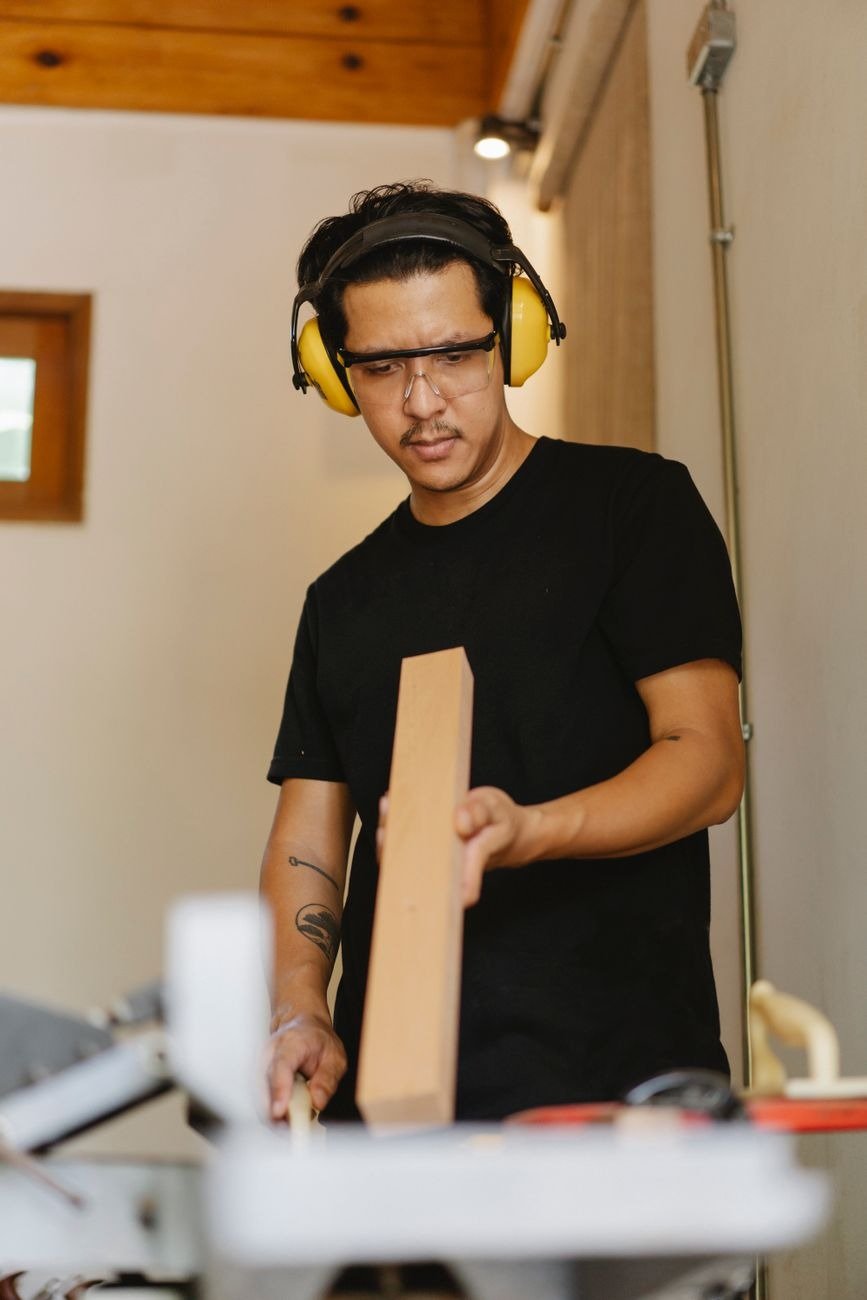 Young concentrated ethnic male carpenter in protective headphones with lumber looking down near professional electric machine in workshop