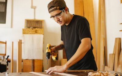 Young attentive ethnic male carpenter in protective glasses with drilling machine and timber in workroom