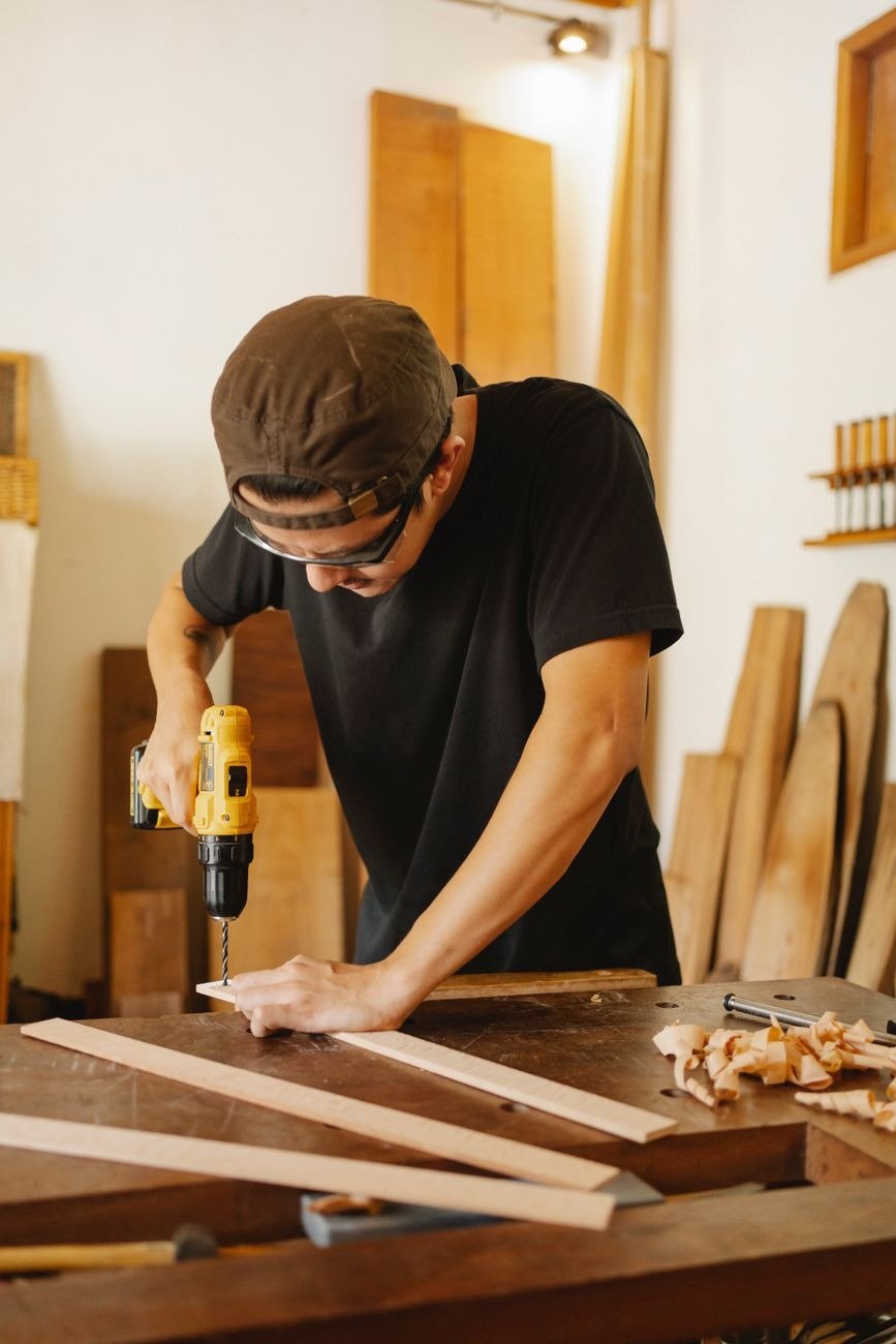 Concentrated craftsman in casual clothes and protective glasses standing near table while working with drill in workshop