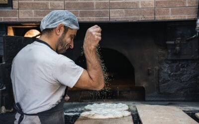 A baker skillfully sprinkles flour on flatbread dough in front of a traditional oven, showcasing baking artistry.