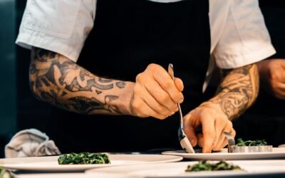 A tattooed chef carefully plates a spinach dish in a professional kitchen setting, showcasing culinary expertise.