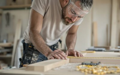 Un menuisier mesure une planche au mètre, un téléphone posé sur l'établi couvert de sciure