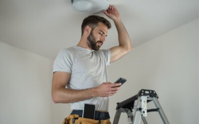 Un électricien sur un escabeau pose un plafonnier, un téléphone à la main