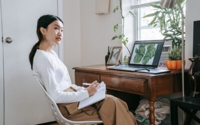 Young woman taking notes at home office with laptop and indoor plants, focused and thoughtful.