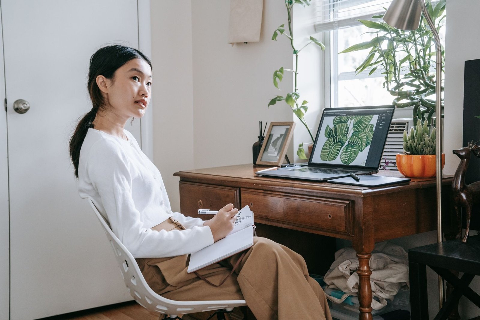 Young woman taking notes at home office with laptop and indoor plants, focused and thoughtful.