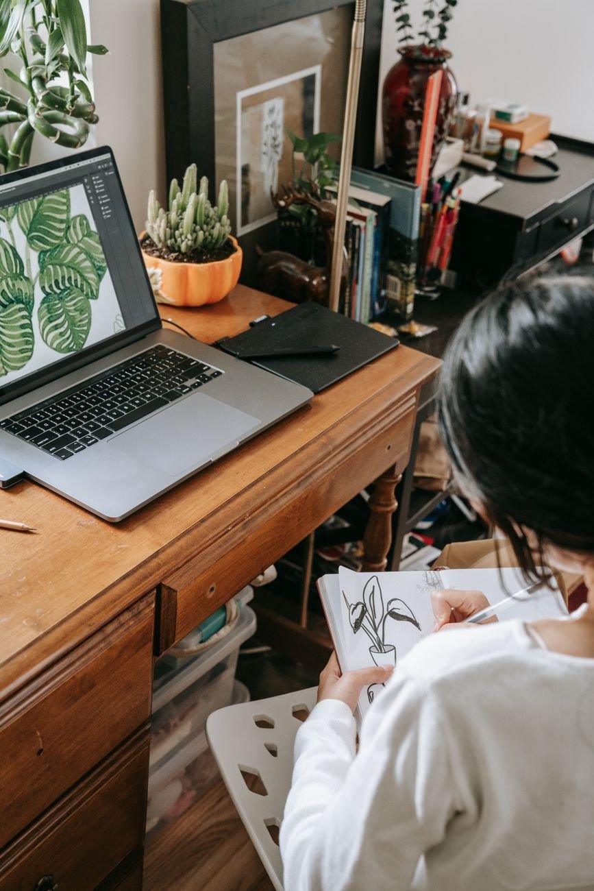 A creative home workspace featuring a laptop and a person sketching plants, with indoor plants on a wooden desk.