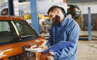 Smiling auto mechanic inspects a car in a service garage, wearing coveralls and holding documents.